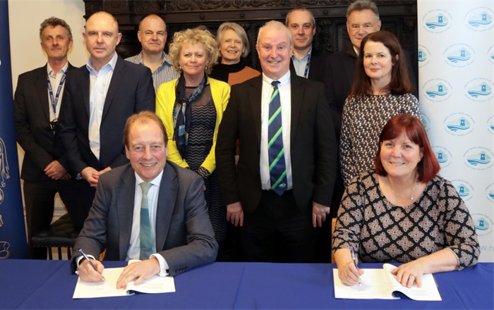 Dyn a dynes yn eistedd wrth y bwrdd yn arwyddo dogfen gyda grŵp o naw o bobl yn sefyll y tu ôl iddyn nhw - i gyd yn edrych ar y camera