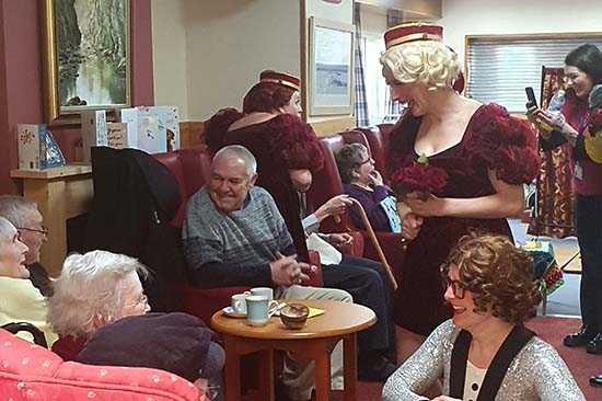 Grŵp o bobl oedrannus yn eistedd i lawr mewn cartref gofal yn siarad â menywod mewn gwisgoedd