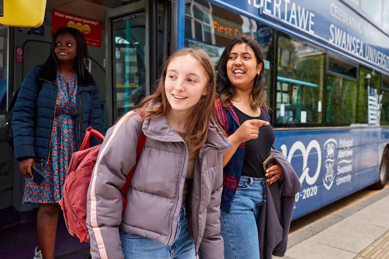 A photo of two students, one male and one female, looking at their phone with a First Cymru bus in the background