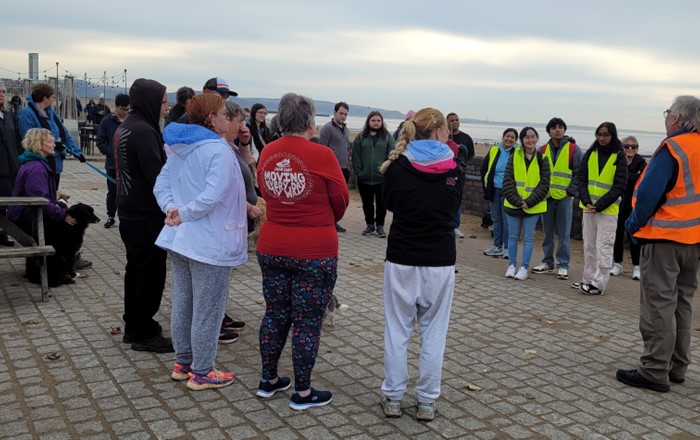 Group of people standing outside on seafront listening to a man in high-vis vest 