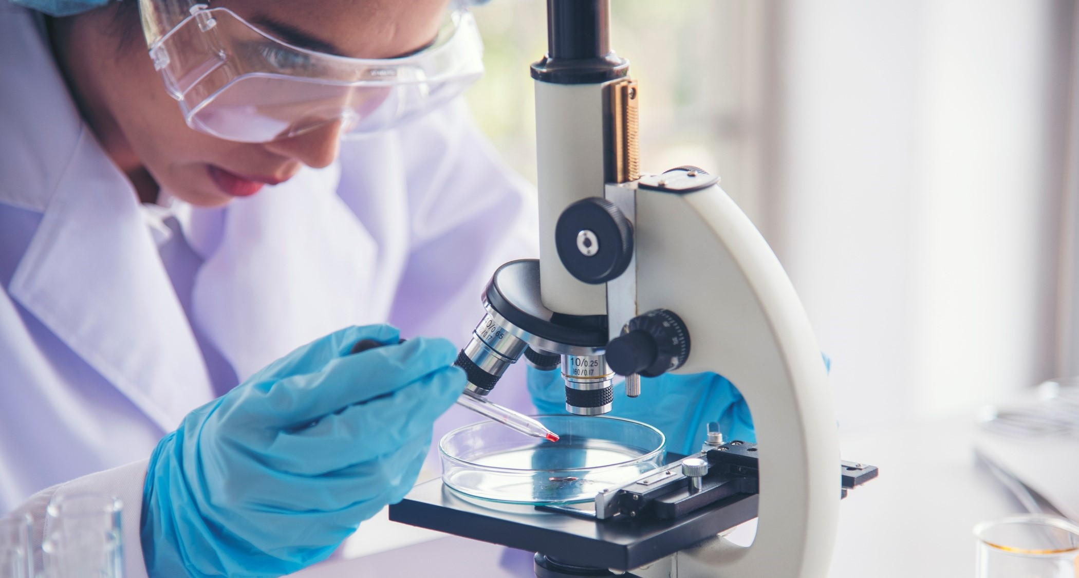 Scientist in a white lab coat and goggles looking through the microscope
